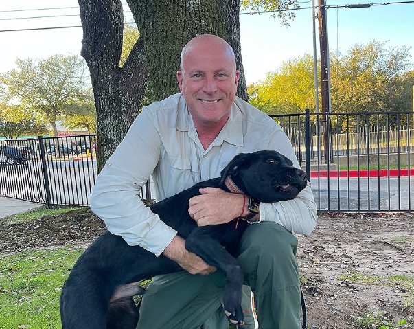Brice Cavanaugh smiling and hugging a black Pitbull in a park with a big oak tree in the background