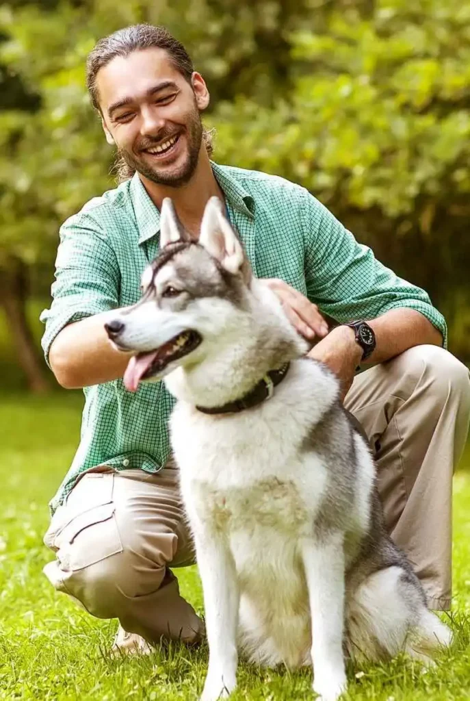 Man in a green shirt smiling and sitting down with his husky in the grass on a sunny day after doing private dog training lessons in Austin tx.